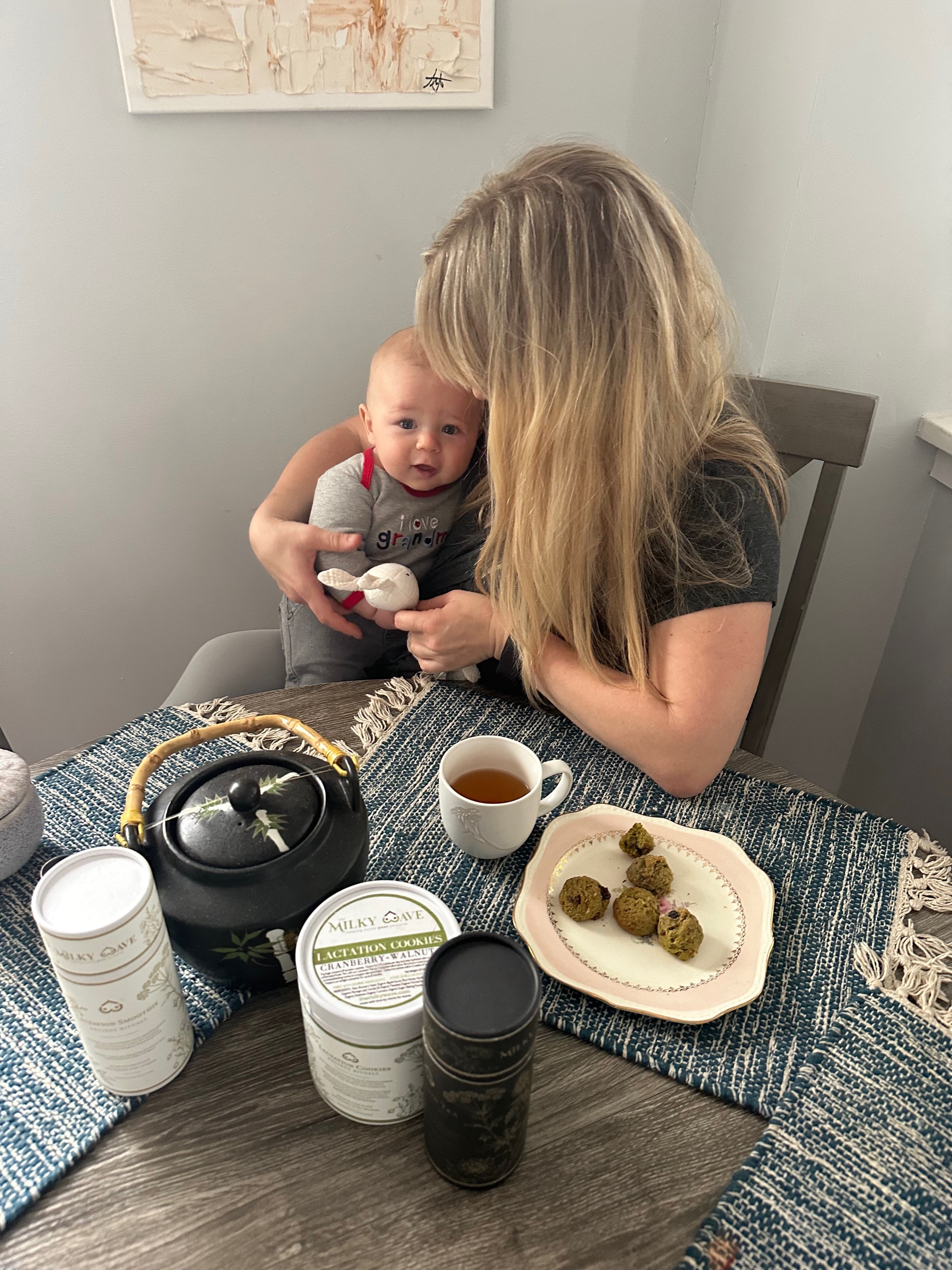 Mom with baby beside The Milky Wave lactation tea and cookies on a table