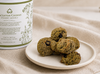 Green cookies on a white plate with a container labeled 'Cranberry Walnut Lactation Cookies' on a beige fabric background